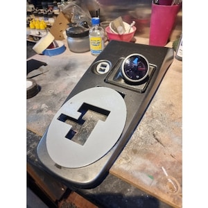 May include: A gray metal panel with a round cutout for a gauge. The gauge is black with a white needle and a white face. The panel has a small, rectangular button on the top right corner.