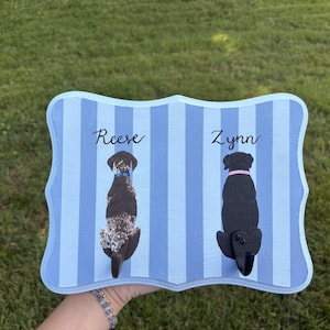 May include: A rectangular wooden wall hook plaque with a light and dark blue vertical striped pattern. It features two painted dog illustrations with the names Reese and Zynn written above them in black script, and two black metal hooks attached.