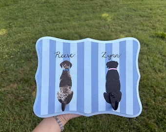 May include: A rectangular wooden wall hook plaque with a light and dark blue vertical striped pattern. It features two painted dog illustrations with the names Reese and Zynn written above them in black script, and two black metal hooks attached.