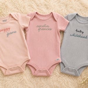 May include: Three ribbed baby bodysuits with short sleeves. The left is light pink with embroidered text "peggy jeane". The center is dusty rose with embroidered text "amelia frances". The right is light blue with embroidered text "baby whitehead".