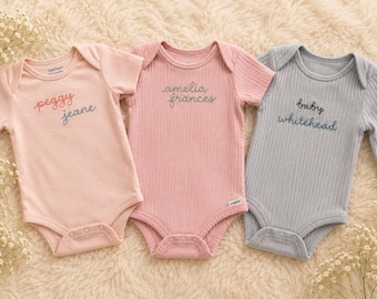 May include: Three ribbed baby bodysuits with short sleeves. The left is light pink with embroidered text "peggy jeane". The center is dusty rose with embroidered text "amelia frances". The right is light blue with embroidered text "baby whitehead".