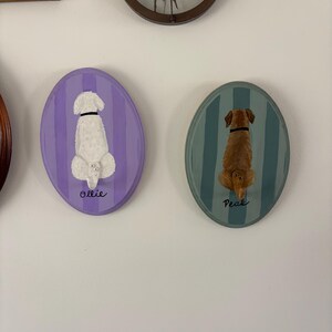 May include: Two oval wall plaques featuring painted dogs from behind. The left plaque is purple with vertical stripes and a white dog labeled Ollie. The right plaque is teal with vertical stripes and a brown dog labeled Peak. Both dogs have collars.