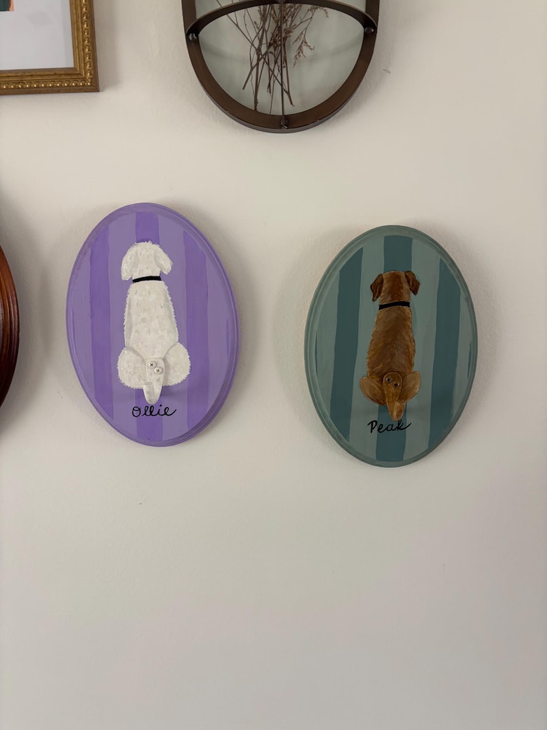 May include: Two oval wall plaques featuring painted dogs from behind. The left plaque is purple with vertical stripes and a white dog labeled Ollie. The right plaque is teal with vertical stripes and a brown dog labeled Peak. Both dogs have collars.