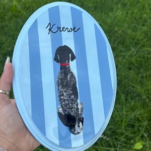 May include: An oval wooden plaque featuring vertical light and dark blue stripes. A painted black and white speckled dog with a red collar is depicted sitting, facing away from the viewer. The name Krewe is written in black script above the dog.