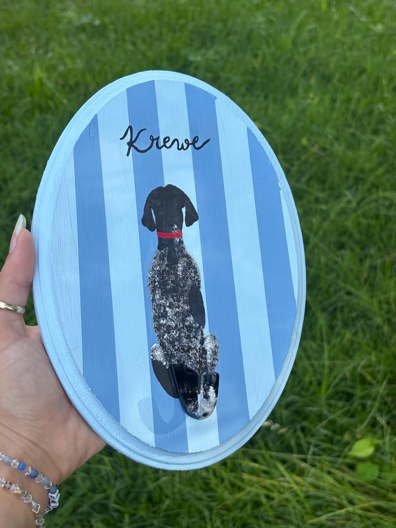 May include: An oval wooden plaque featuring vertical light and dark blue stripes. A painted black and white speckled dog with a red collar is depicted sitting, facing away from the viewer. The name Krewe is written in black script above the dog.
