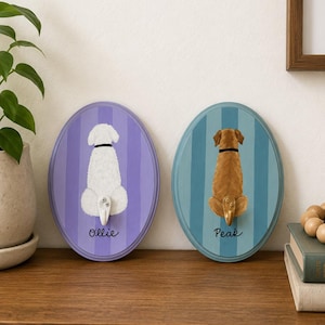 May include: Two oval wall hooks featuring painted dog backsides. The left hook is purple with a white dog and the name Ollie. The right hook is blue with a brown dog and the name Peak. Each dog has a small hook attached at the tail position.