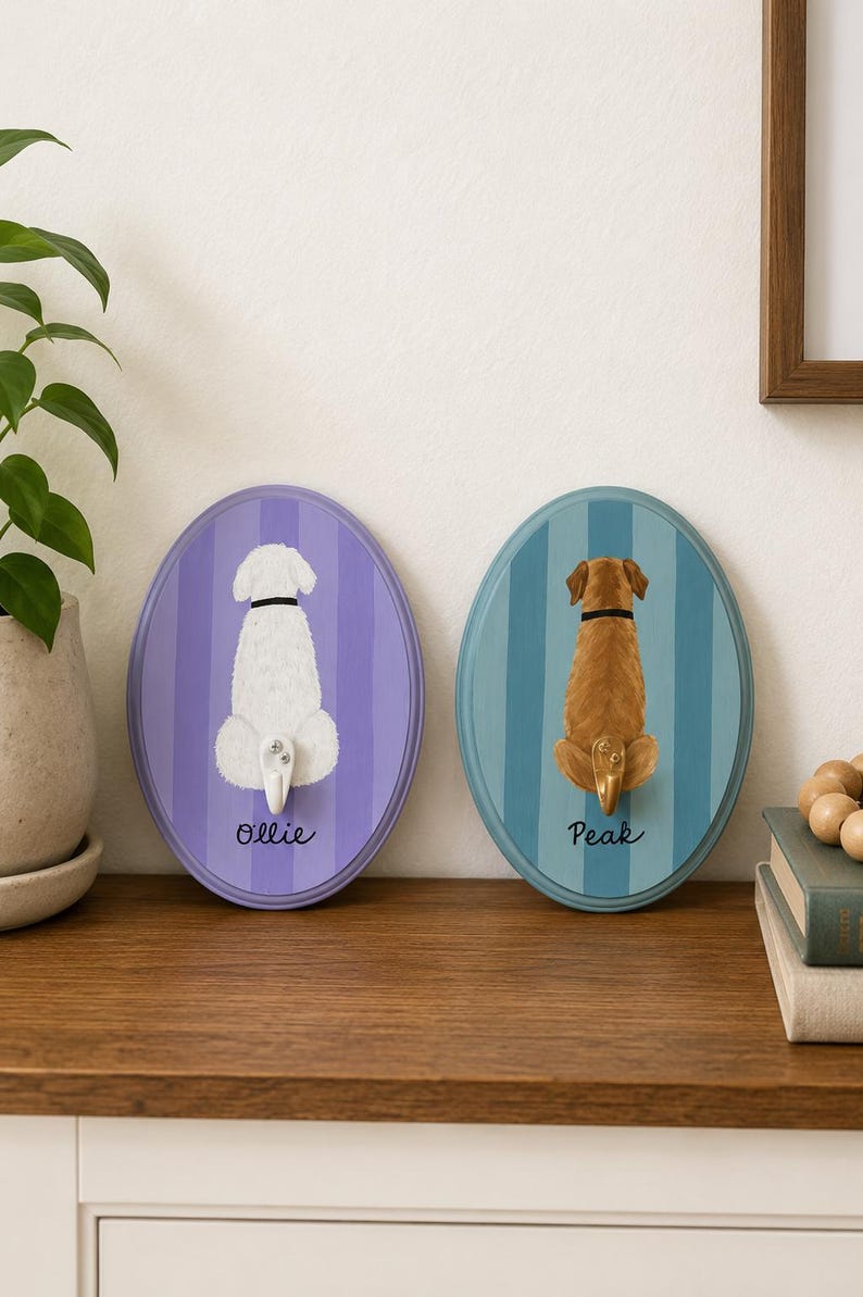 May include: Two oval wall hooks featuring painted dog backsides. The left hook is purple with a white dog and the name Ollie. The right hook is blue with a brown dog and the name Peak. Each dog has a small hook attached at the tail position.