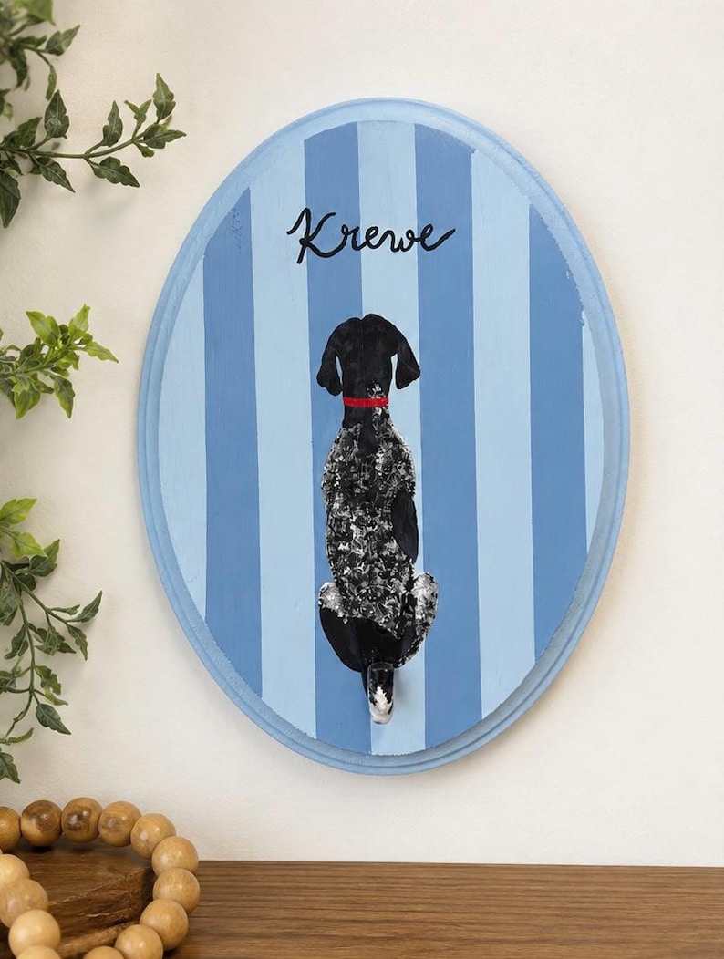 May include: An oval wooden wall plaque featuring a painted black and white speckled dog viewed from behind, wearing a red collar. The background consists of vertical light and medium blue stripes, with the word Krewe written in black script.