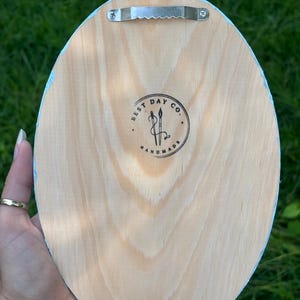 May include: An oval-shaped light-colored wooden plaque with a metal sawtooth hanger attached to the top. The center features a circular black ink stamp with the text BEST DAY CO. HANDMADE and an illustration of two crossed paintbrushes.