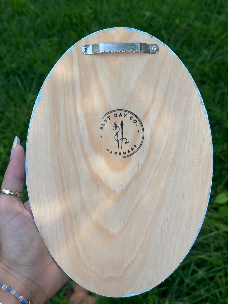May include: An oval-shaped light-colored wooden plaque with a metal sawtooth hanger attached to the top. The center features a circular black ink stamp with the text BEST DAY CO. HANDMADE and an illustration of two crossed paintbrushes.