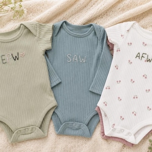May include: Three ribbed baby bodysuits: a sage green one with embroidered 'EPW' and a heart, a blue long-sleeve one with 'SAW' embroidered in white, and a white one with a small pink floral pattern and the embroidered letters 'AFW'.