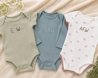 May include: Three ribbed baby bodysuits: a sage green one with embroidered 'EPW' and a heart, a blue long-sleeve one with 'SAW' embroidered in white, and a white one with a small pink floral pattern and the embroidered letters 'AFW'.