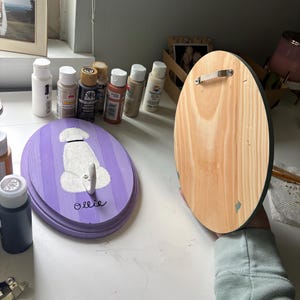 May include: Two oval wooden plaques, one painted with purple stripes and a white dog silhouette with the name Ollie written in black, and the other showing the natural wood grain back with a metal sawtooth hanger attached for wall mounting.