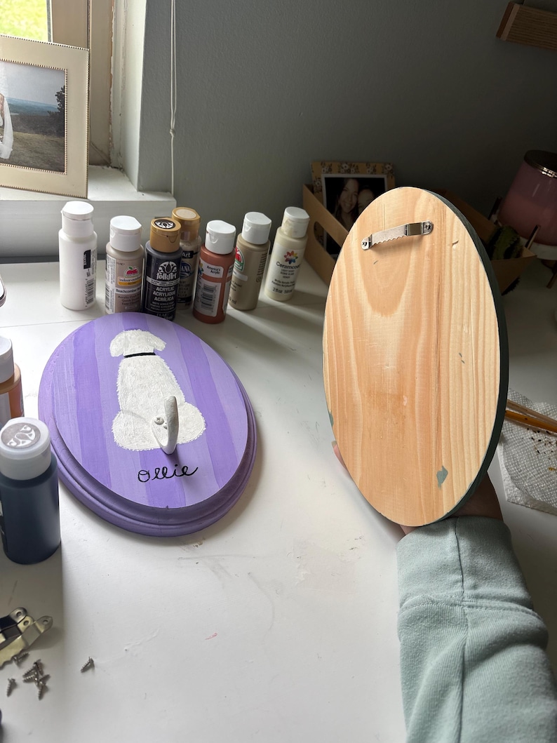 May include: Two oval wooden plaques, one painted with purple stripes and a white dog silhouette with the name Ollie written in black, and the other showing the natural wood grain back with a metal sawtooth hanger attached for wall mounting.