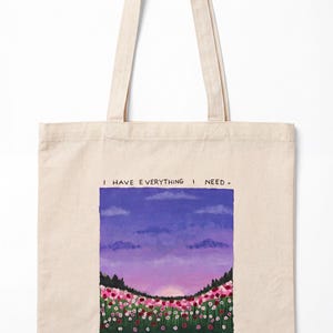 May include: A beige canvas tote bag featuring a printed landscape illustration of a field of pink and red flowers under a purple sunset sky. The text "I HAVE EVERYTHING I NEED" is printed above the scene, and "psalm 23:1" is printed below.