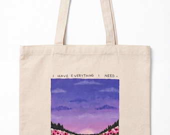May include: A beige canvas tote bag featuring a printed landscape illustration of a field of pink and red flowers under a purple sunset sky. The text "I HAVE EVERYTHING I NEED" is printed above the scene, and "psalm 23:1" is printed below.