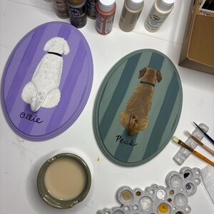 May include: Two oval wooden plaques featuring hand-painted dogs viewed from behind. One plaque is purple with a white dog named Ollie, the other is teal with a brown dog named Peak. Each plaque includes a small metal hook. Acrylic paints and brushes are nearby.