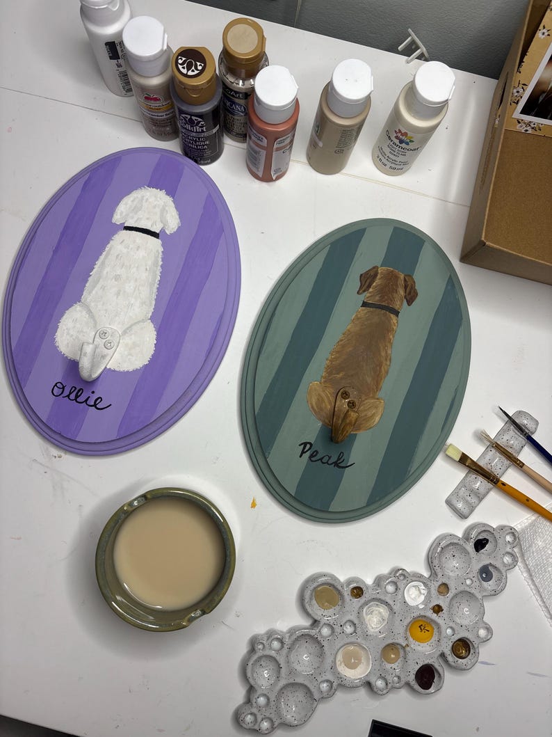 May include: Two oval wooden plaques featuring hand-painted dogs viewed from behind. One plaque is purple with a white dog named Ollie, the other is teal with a brown dog named Peak. Each plaque includes a small metal hook. Acrylic paints and brushes are nearby.