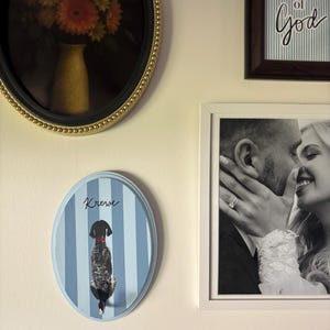 May include: An oval wall plaque featuring a light blue and white vertical striped background with a black dog illustration wearing a red collar. The word Krew is written in black script above the dog. The plaque has a light blue painted border.