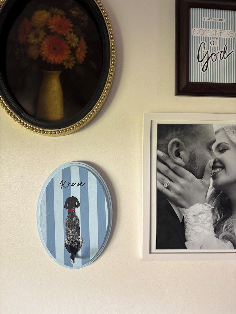 May include: An oval wall plaque featuring a light blue and white vertical striped background with a black dog illustration wearing a red collar. The word Krew is written in black script above the dog. The plaque has a light blue painted border.