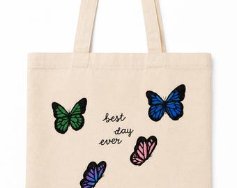 May include: A beige canvas tote bag featuring six colorful butterfly illustrations in green, blue, pink, purple, light blue, and orange. The text "best day ever" is printed in black cursive lettering in the center of the bag between the butterflies.