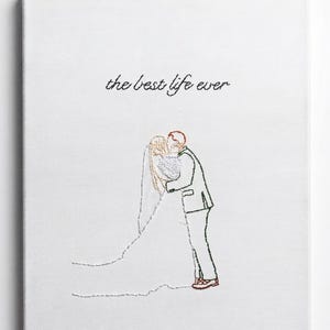 May include: A white canvas featuring black thread embroidery of a couple kissing, with the bride in a long veil and the groom in a suit. The text 'the best life ever' is embroidered in black cursive script above the couple on the white fabric.