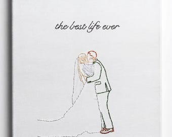 May include: A white canvas featuring black thread embroidery of a couple kissing, with the bride in a long veil and the groom in a suit. The text 'the best life ever' is embroidered in black cursive script above the couple on the white fabric.
