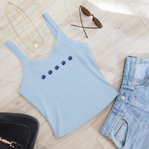 May include: A light blue tank top with dark blue dot pattern, sunglasses, gold necklace, black purse, and denim shorts. The casual outfit is laid out on a light wood surface.