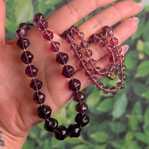May include: A vintage necklace with dark purple, faceted beads. The beads are strung together, forming a necklace with a clasp. The necklace is held in a hand against a green backdrop.