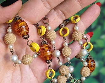 May include: A vintage necklace featuring brown and yellow glass beads, white pearl beads, and yellow plastic beads. The necklace is on a brown chain.