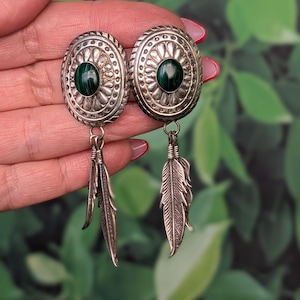 May include: A pair of oval-shaped silver earrings featuring a central dark green malachite stone surrounded by embossed radial patterns. Each earring includes two dangling silver feather charms attached to the base by small metal jump rings.