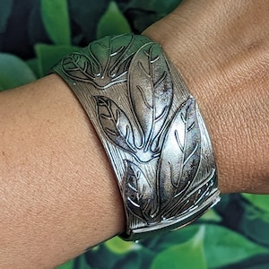 Puede incluir: Un brazalete de metal plateado con un diseño de hojas. El brazalete es ancho y tiene una superficie texturizada.