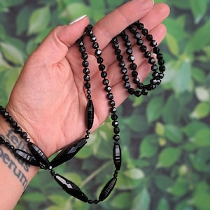 May include: A black beaded necklace featuring faceted and elongated beads. The necklace is displayed against a green, leafy backdrop, highlighting its dark hue and detailed design. The necklace is a piece of jewellery.