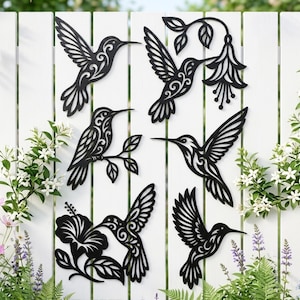 Könnte beinhalten: Schwarze Metall-Wanddekorationen in Form von Kolibris, mit detaillierten Designs, an einem weißen Zaun befestigt. Einige Stücke zeigen Blumen. Ideal für die Innen- oder Außendekoration.