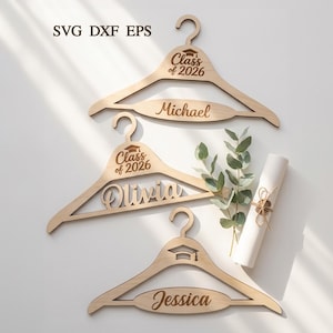May include: Wooden graduation hangers with names and "Class of 2026" text. The hangers are light brown and feature a graduation cap design. One hanger has the name "Michael", another "Olivia", and the third "Jessica". A rolled diploma and greenery are also present.