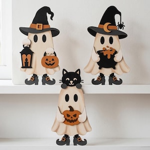 May include: Three wooden ghost figurines featuring Halloween themes. One ghost wears a witch hat and holds a lantern and pumpkin. Another wears a witch hat with a moon and spider, holding a cauldron. The third ghost has a black cat on its head.