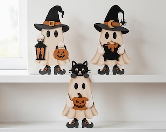 May include: Three wooden ghost figurines featuring Halloween themes. One ghost wears a witch hat and holds a lantern and pumpkin. Another wears a witch hat with a moon and spider, holding a cauldron. The third ghost has a black cat on its head.