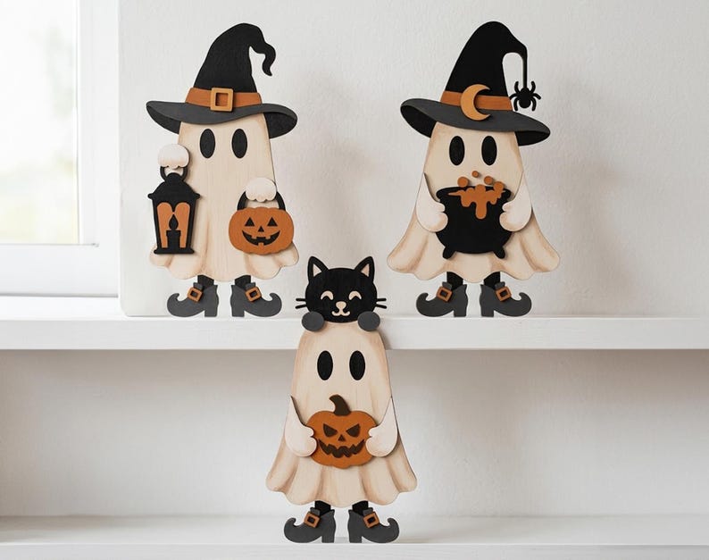 May include: Three wooden ghost figurines featuring Halloween themes. One ghost wears a witch hat and holds a lantern and pumpkin. Another wears a witch hat with a moon and spider, holding a cauldron. The third ghost has a black cat on its head.