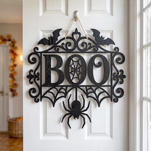 Puede incluir: Un letrero de pared de madera negra con la palabra BOO en letras may&uacute;sculas grandes, con un patr&oacute;n de telara&ntilde;a dentro de la letra O. El letrero est&aacute; decorado con dos murci&eacute;lagos en la parte superior, volutas y una ara&ntilde;a colgante.