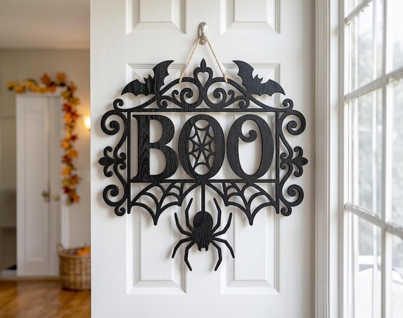 Puede incluir: Un letrero de pared de madera negra con la palabra BOO en letras may&uacute;sculas grandes, con un patr&oacute;n de telara&ntilde;a dentro de la letra O. El letrero est&aacute; decorado con dos murci&eacute;lagos en la parte superior, volutas y una ara&ntilde;a colgante.
