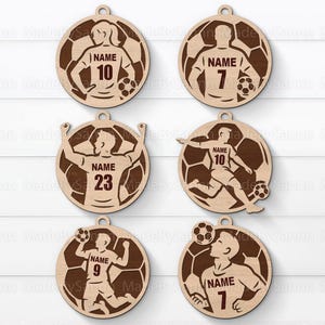 May include: Six wooden soccer-themed ornaments. Each ornament features a soccer player design with a jersey number and the word "NAME". The ornaments are circular with a soccer ball background and a hole for hanging.