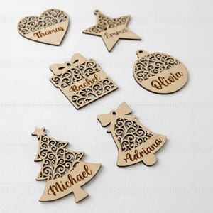Può includere: Una collezione di decorazioni natalizie in legno, tra cui un cuore, una stella, un pacco regalo, una palla, un albero di Natale e una campana. Ogni ornamento presenta un motivo traforato decorativo e un nome. Realizzati in legno chiaro.