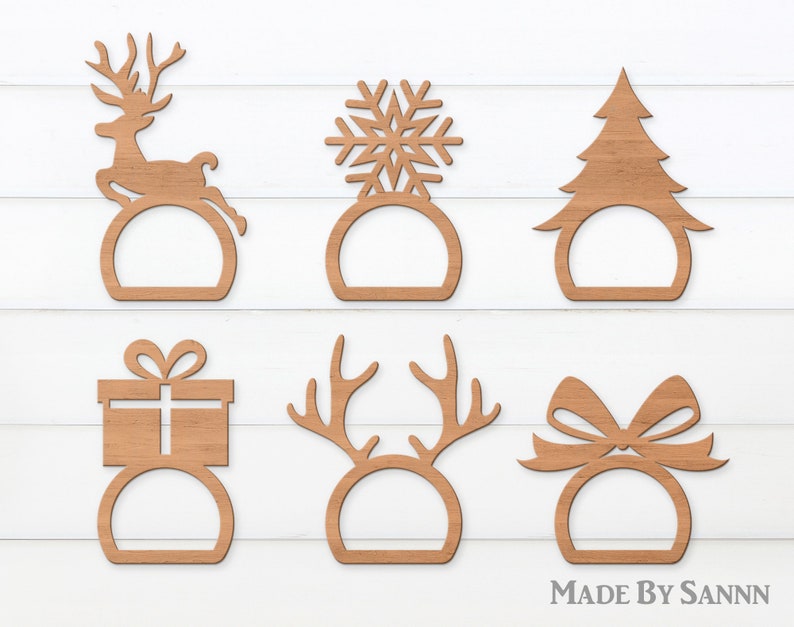 May include: Six wooden Christmas-themed napkin rings in various designs: a reindeer, snowflake, Christmas tree, gift box, antlers, and a bow. The napkin rings are made of light brown wood and are displayed on a white background.