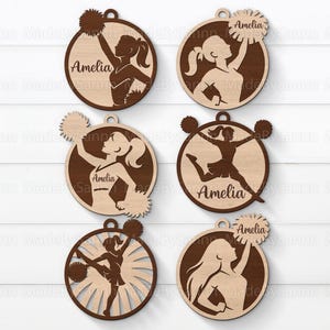 May include: A collection of wooden ornaments depicting cheerleader figures in action poses, set within circular frames. Each ornament is engraved with the name "Amelia". The ornaments are crafted from light and dark brown wood.