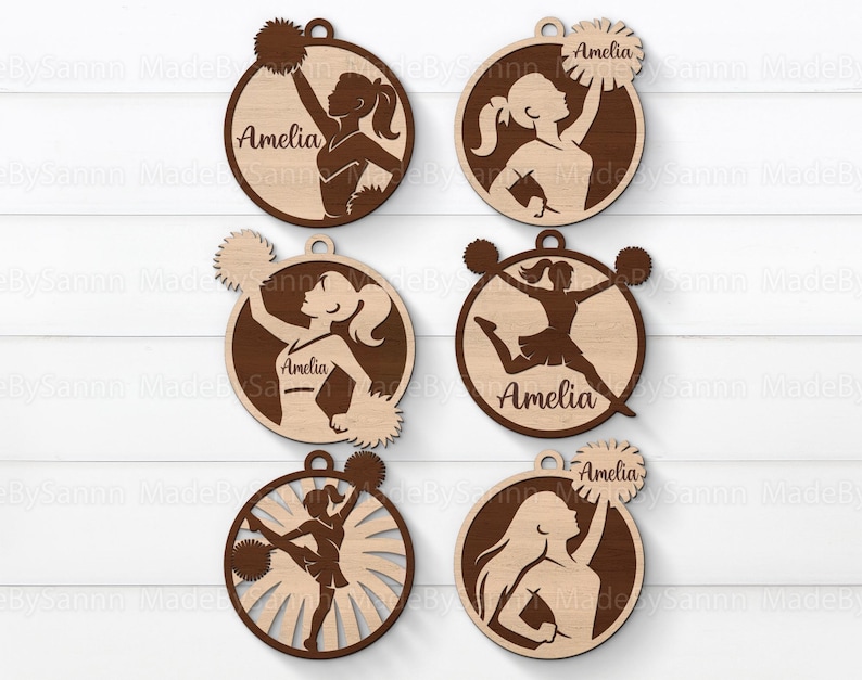May include: Wooden ornaments featuring cheerleader silhouettes in various poses, each within a circular frame. The name "Amelia" is inscribed on each ornament. The ornaments are made of light and dark brown wood.