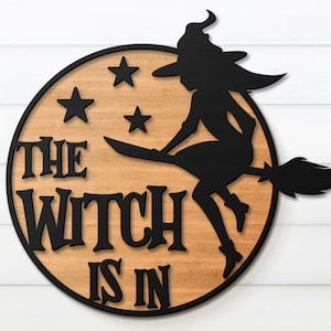 K&ouml;nnte beinhalten: Ein rundes Holzschild mit dem Text "The Witch Is In" in schwarzen Buchstaben. Eine schwarze Silhouette einer Hexe, die auf einem Besen reitet, mit Sternen im Hintergrund.