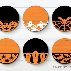 Puede incluir: Seis letreros redondos de madera con un diseño dividido. La mitad superior de cada letrero es naranja y la mitad inferior es negra. Cada letrero presenta un diseño diferente con temática de Halloween, que incluye calabazas, fantasmas, murciélagos, arañas y lápidas. El texto "MADE BY SANNN" es visible en la parte inferior de la imagen.