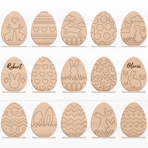 May include: Set of 12 wooden Easter egg shapes with various designs, including hearts, butterflies, bunnies, and geometric patterns. Some eggs have names like "Robert" and "Olivia" engraved on them.