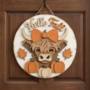 May include: A round wooden wall sign featuring a layered Highland cow illustration with an orange bow, three pumpkins, and two maple leaves. The text "Hello Fall" is displayed in raised wooden lettering at the top, suspended by a jute rope.