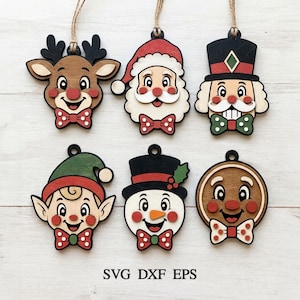 May include: Six wooden Christmas ornaments featuring cartoon faces of a reindeer, Santa Claus, a nutcracker, an elf, a snowman, and a gingerbread cookie, each with a small hanging loop and the text SVG DXF EPS printed at the bottom of the image.